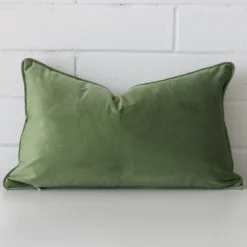 Sage cushion leans elegantly against a brick wall. It has been crafted from a high quality velvet material and has a rectangle shape.