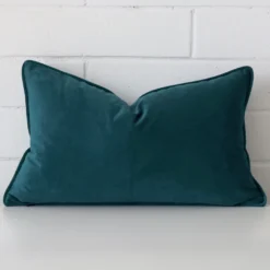 Teal cushion positioned in front of a brick wall. It has rectangle dimensions and is made from a velvet material.